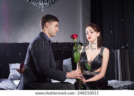 Close up Young Man Offering Rose Flower to Sad Girlfriend While Holding a Jewelry Box Behind his Back. Captured at the Bedroom.
