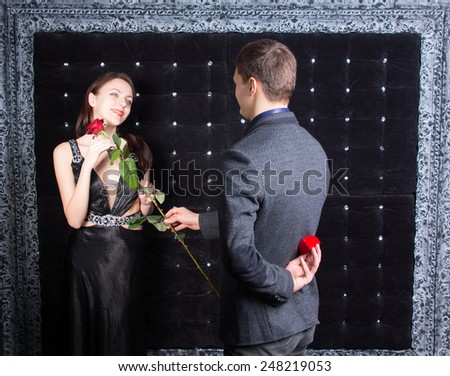 Romantic young man asking a gorgeous young woman in an elegant black cocktail dress to marry him presenting her with a rose before producing the ring