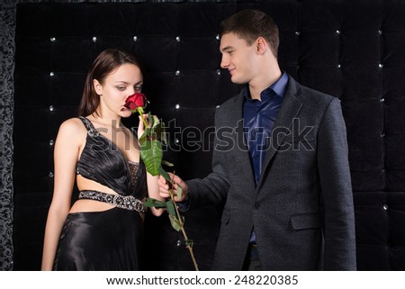 Close up Pretty Young Woman in Sexy Black Dress Smelling the Red Rose Flower as her Handsome Man Holding it. Captured with Sparkling Black Background.