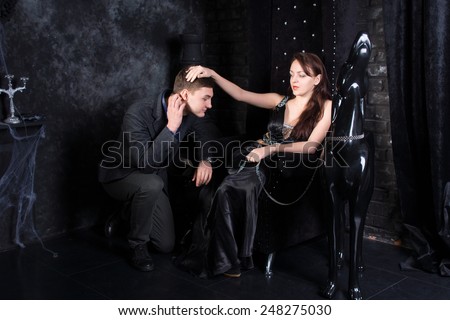 Woman Wearing Formal Black Dress Sitting on Throne with Man Kneeling and Dog Statue Beside Her