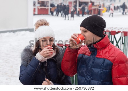 Young couple standing outdoors in an urban square in winter snow sipping hot drinks on a cold day from takeaway cups, people walking in the distance