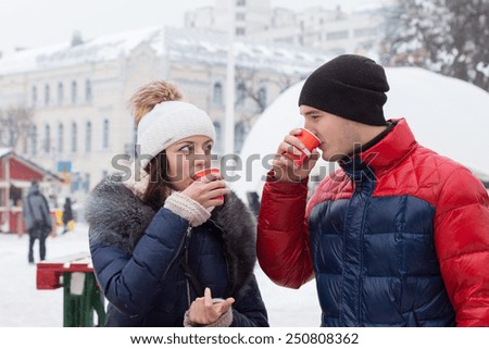 Young couple in warm winter clothing, scarves and knitted caps standing sipping hot drinks in a wintry town square