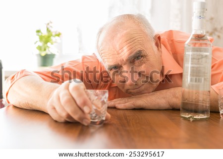 Close up Drunk Elderly Holding Small Shot Glass at the Wooden Table with Vodka While Looking at the Camera.