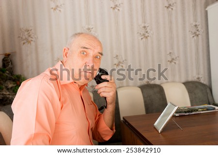 Close up Surprised Old Man Shaving his Beard Using Electric Razor at his Table Inside his House While Looking at the Camera.
