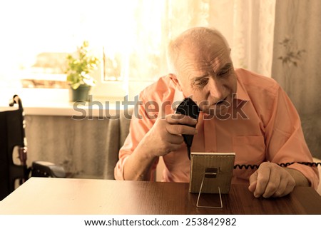 Close up Serious Senior Man Shaving Beard with Electric Razor at the Wooden Table with Small Mirror in the Living Room