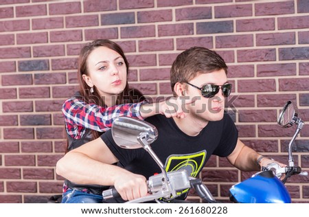 Young Couple on Motorcycle in front of Brick Wall with Woman Pointing Forward Over Mans Shoulder