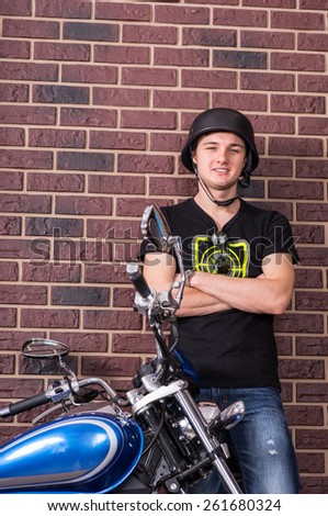 Close Up Portrait of Smiling Young Man Wearing Helmet Standing with Arms Crossed Against Brick Wall Next to Blue Motorcycle