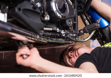 Young man doing maintenance on his motorbike lying on the floor underneath with a spanner in his hand, close up view