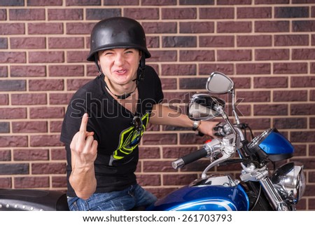 Young thug sitting on his blue motorbike making a rude derisive gesture with his middle finger with a provocative expression