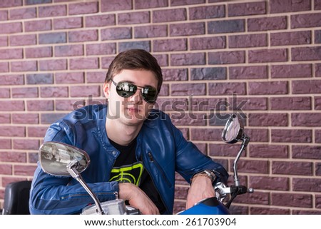 Trendy young man on a motorbike leaning over the handlebars in his fashionable sunglasses to smile at the camera