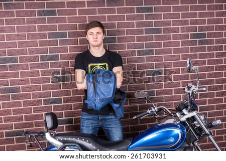 Young Handsome Man Standing with his Jacket and Motorcycle in Front of a Brick Wall While Looking at the Camera.