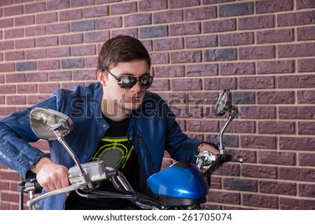 Close up Handsome Young Man Wearing Sunglasses and Jacket on a Sports Motorbike with Brick Wall Background.