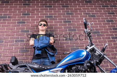 Handsome Young Man with Sunglasses Holding his Jacket While Standing Behind his Blue Motorbike on a Brick Wall Background.