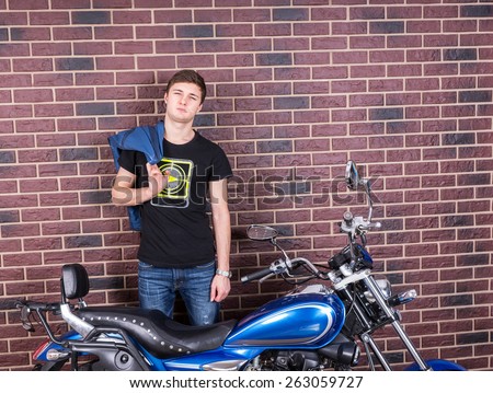 Young Handsome Man, Holding his Jacket Over his Shoulder, Standing In Front of a Brick Wall with his Blue Custom Motorcycle