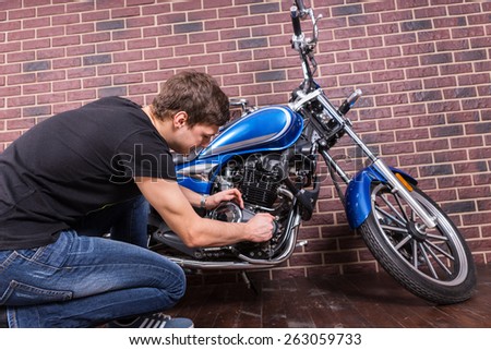 Sitting Young Handsome Guy in Casual Outfit Repairing his Blue Motorcycle Manually in Front of House Brick Wall