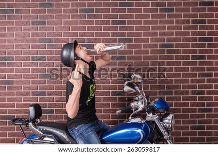 Hyped up young man sitting on his motorbike in his helmet drinking alcohol directly from the bottle and punching the air with his fist