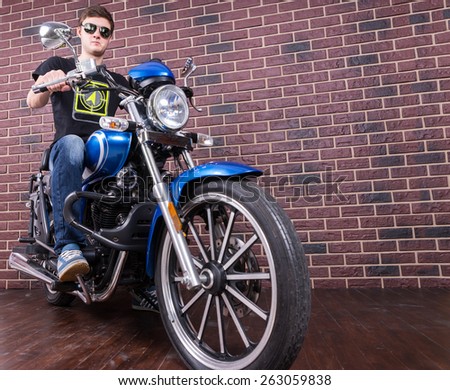 Low Angle Full Length Portrait of Young Man Sitting on Classic Blue Motorcycle in front of Brick Wall