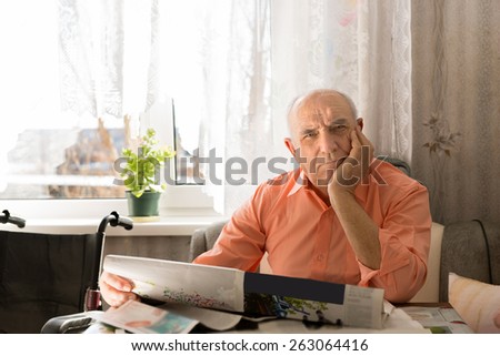Close up Sitting Old Man Holding Newspaper While Looking at the Camera with One Hand on the Face.