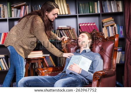 Librarian waking a sleeping man in the library sitting in an armchair with a book on his chest leaning over and touching him on the shoulder