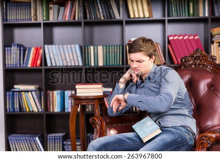 Serious Young Guy in Trendy Fashion Sitting and Waiting at Library Chair with a Book, Looking at his Wristwatch