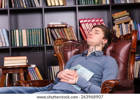 Tired young man asleep in the library reclining in a comfortable leather wooden carved armchair with his open book resting on his stomach