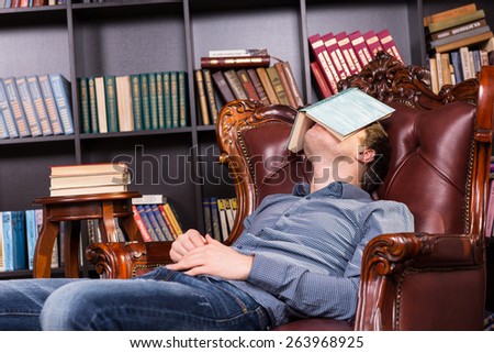 Exhausted young man sleeping in a library relaxing in a comfortable leather armchair with an open book over his eyes