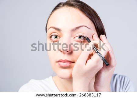Close up Smiling Young Woman Applying Eyeliner Makeup to Left Eye While Looking at the Camera on a Gray Background.