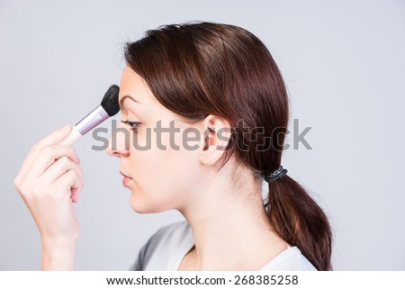 Close up Young Pretty Woman in Side View Applying Makeup on Forehead Using Brush While Looking Down, Captured in Studio with Gray Background.