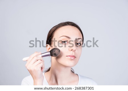 Close up Pretty Young Woman Applying Foundation Makeup on her Face Using a Brush While Looking at the Camera, Isolated on a Gray Background.