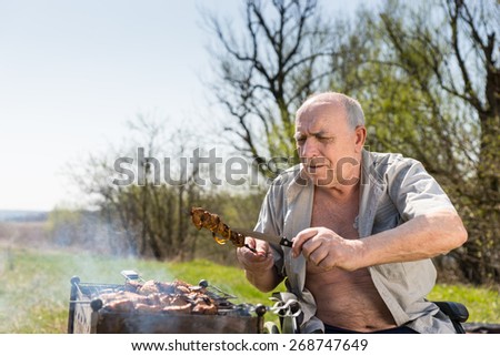 Sitting Old Man Checking if Grilled Meat on a Stick are Well-Cooked While Doing a Barbecue at the Park.