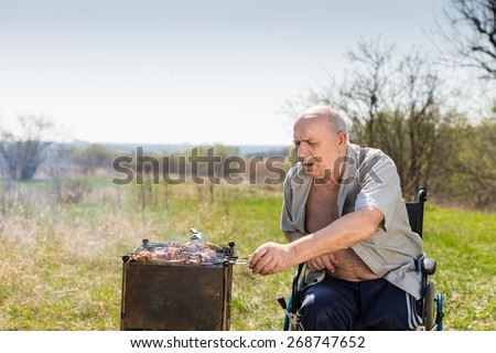 Disabled Elderly Man with Unbuttoned Shirt Sitting on his Wheelchair While Grilling Some Meat Sausages to Eat at the Park Alone on a Hot Morning.