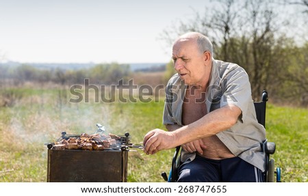 Aged Man with Unbuttoned Shirt Sitting on his Wheelchair While Grilling for his Lunch Under the Heat of the Sun at the Park.