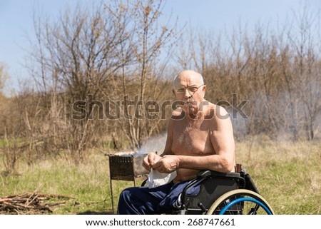 Topless Handicap Senior Man, Camping at the Park, Sitting on his Wheelchair and Preparing Something While Looking at the Camera.