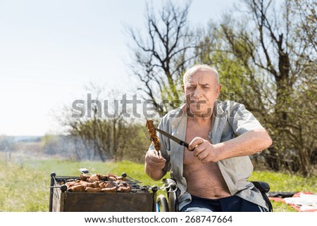 Elderly Man with Unbuttoned Shirt and Sitting on his Wheelchair Holding Grilled Meat on Stick and Knife and Looking at the Camera While Grilling at the Park.