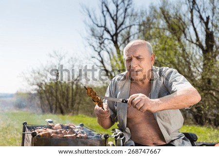 Happy Old Man with Open Shirt and Sitting on his Wheelchair Checking the Grilled Meat on Stick Using Knife to Know if Properly Cooked.