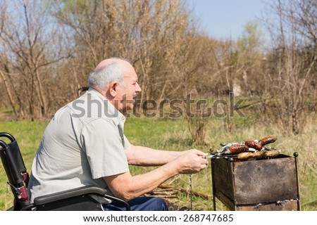Male Elderly Sitting on his Wheelchair Preparing Grilled Food to Eat Outside on a Tropical Climate. Captured in Side View.