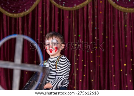 Young Boy Dressed as Clown Holding Large Double Barreled Gun with Iron Sight, Standing on Stage with Red Curtain