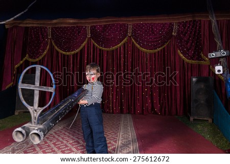 Young Boy Dressed as Clown Standing on Stage Holding Large Double Barreled Shot Gun with Iron Sight, Perspective Exaggerates Gun Size