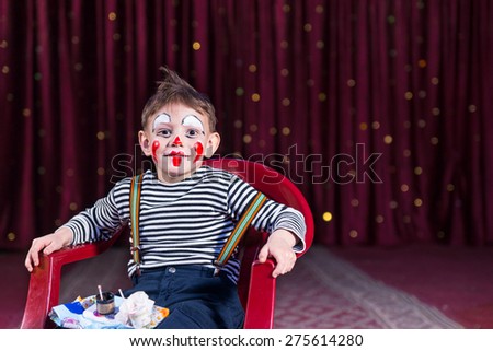 Portrait of Boy Wearing Clown Make Up Sitting in Red Plastic Chair with Tray of Make Up on Stage with Red Curtain, Copy Space to the Right