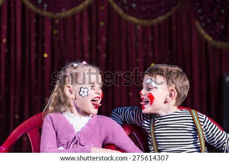 Boy and Girl Wearing Clown Make Up Sitting in Chairs Side by Side and Sticking Tongues Out at Each Other
