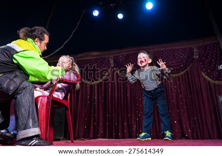 Man Applying Make Up to Face of Blond Girl Sitting in Chair While Young Boy Clown Performs on Stage in Background