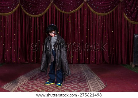 Young Boy Wearing Clown Make Up Standing on Stage in Over Sized Leather Jacket with Red Curtain in Background