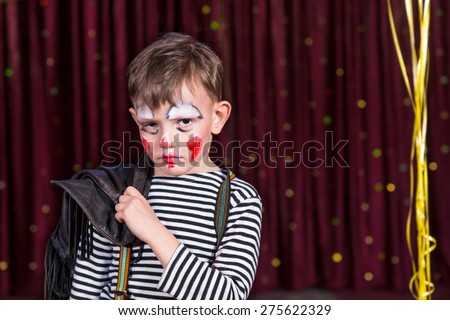 Sulky little boy wearing face paint on stage looking at the camera with a soulful sad expression as he stands in front of the curtain