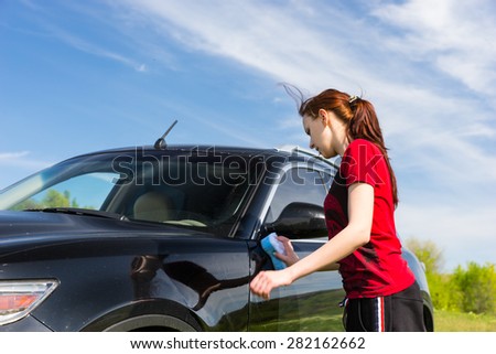 Young Woman with Long Auburn Hair and Wearing Red Shirt Washing Black Luxury Vehicle with Sponge in Green Field on Sunny Day