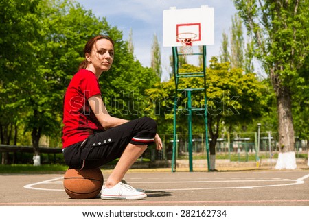 Full Length of Competitive Looking Young Woman Sitting on Top of Basketball on Court in Lush Green Park with Backboard and Basket in Background