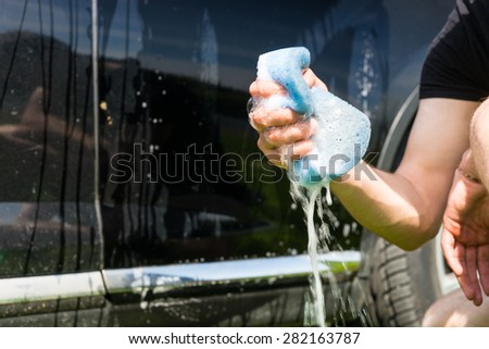 Close Up of Man Washing Car, Crouching Next to Black Car and Squeezing Soapy Blue Sponge Outdoors