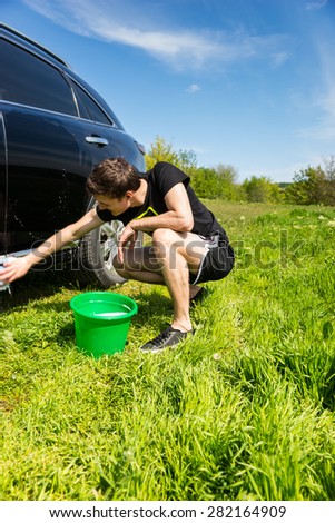 Man Washing Car with Soapy Sponge, Crouching Next to Green Bucket in Green Grassy Field on Bright Sunny Day with Blue Sky