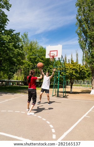 Young Athletic Couple Playing Basketball Together, Man in front of Net Blocking Womans Shot on Outdoor Court in Lush Green Park