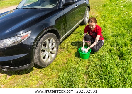 High Angle View of Woman with Green Bucket Wringing Out Soapy Sponge and Washing Black Luxury Vehicle in Green Field on Bright Sunny Day with Blue Sky