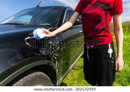 Close Up of Mid Section of Woman Wearing Athletic Clothing Washing Black Luxury Car with Soapy Sponge in Green Field on Bright Sunny Day